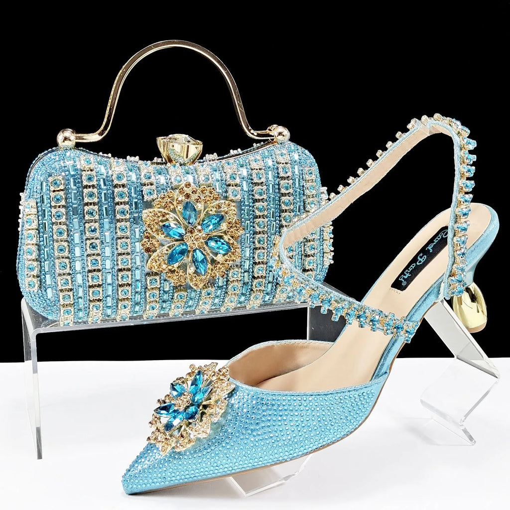Women's black high heel sandal with rhinestone flower decoration and ankle chain, shown with matching clutch bag
