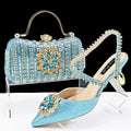 Women's black high heel sandal with rhinestone flower decoration and ankle chain, shown with matching clutch bag