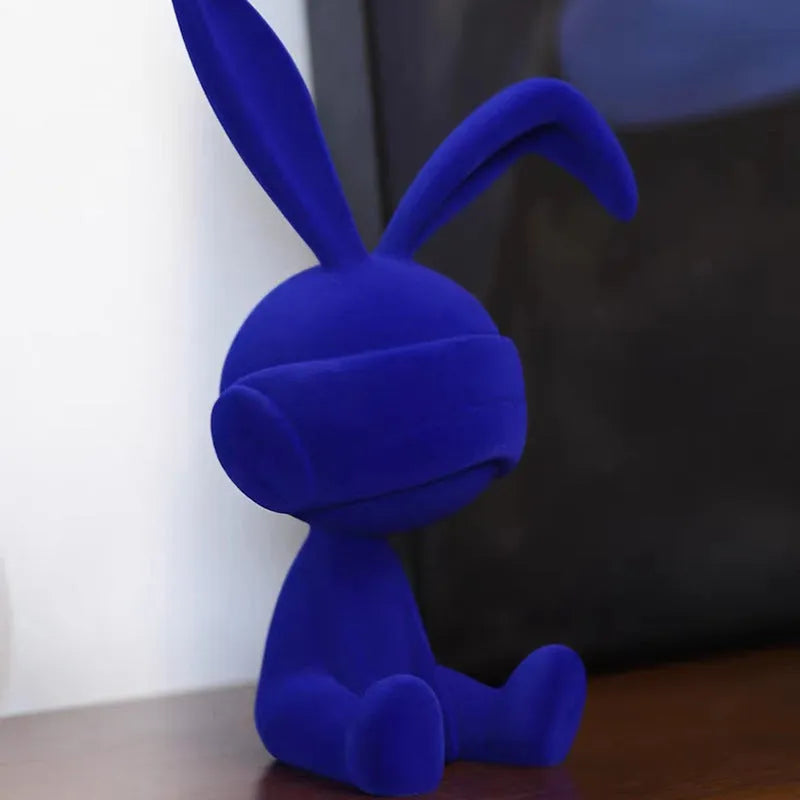 Resin table ornament of a rabbit wearing a suede blindfold with ribbon and tassel detail.