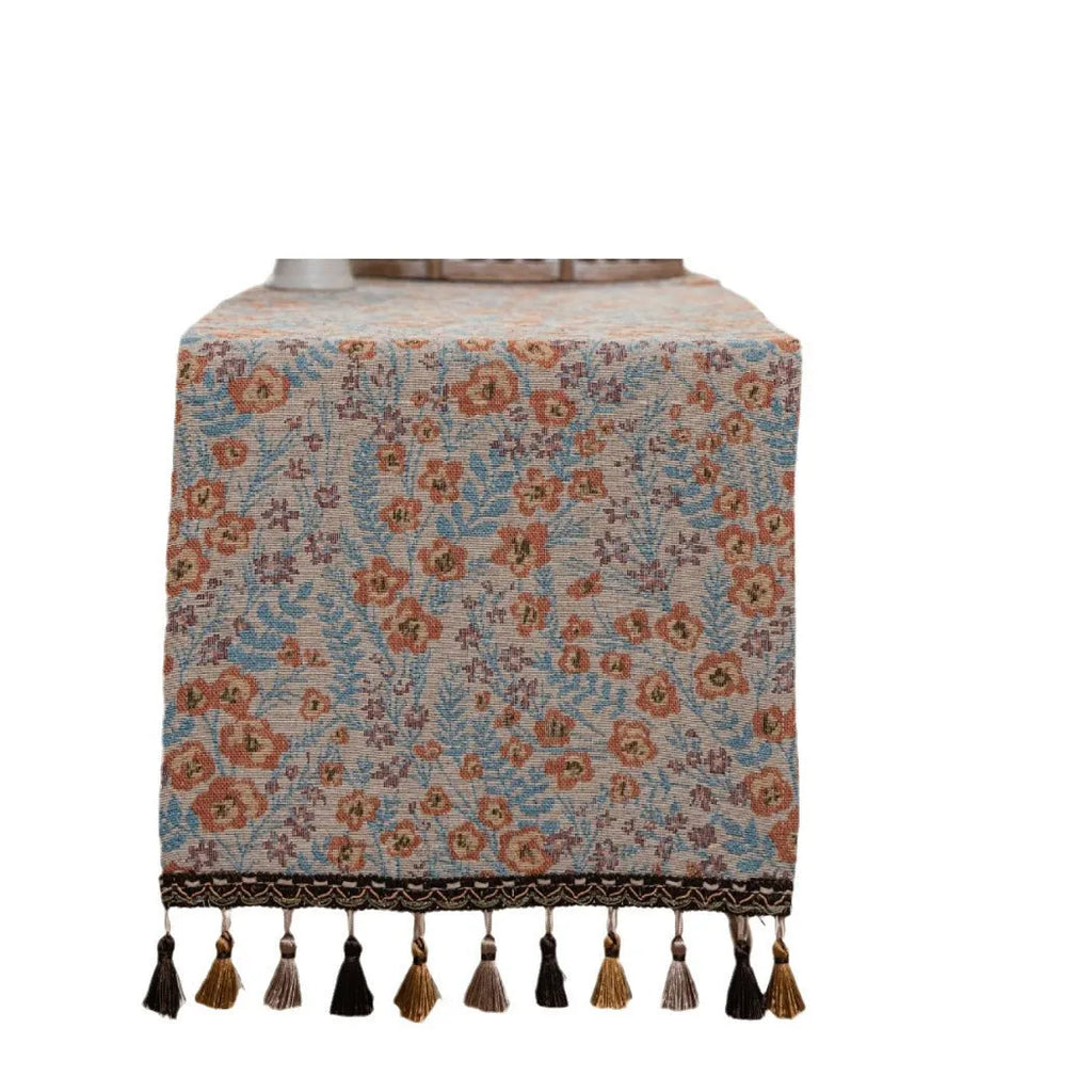 Long beige table runner with floral Jacquard pattern and tassels, shown on a wooden table.