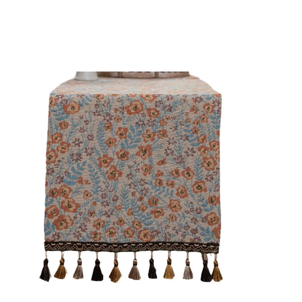 Long beige table runner with floral Jacquard pattern and tassels, shown on a wooden table.