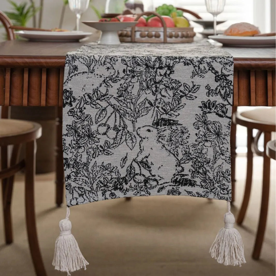 Long beige table runner with floral Jacquard pattern and tassels, shown on a wooden table.
