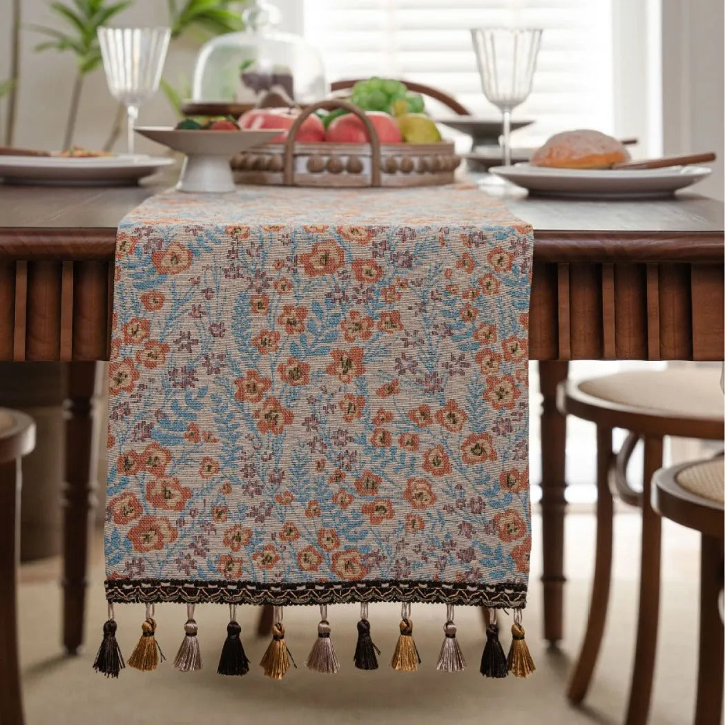 Long beige table runner with floral Jacquard pattern and tassels, shown on a wooden table.