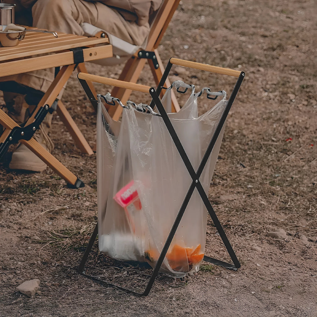 Black foldable outdoor iron trash rack with wooden handle holding a white bin bag.