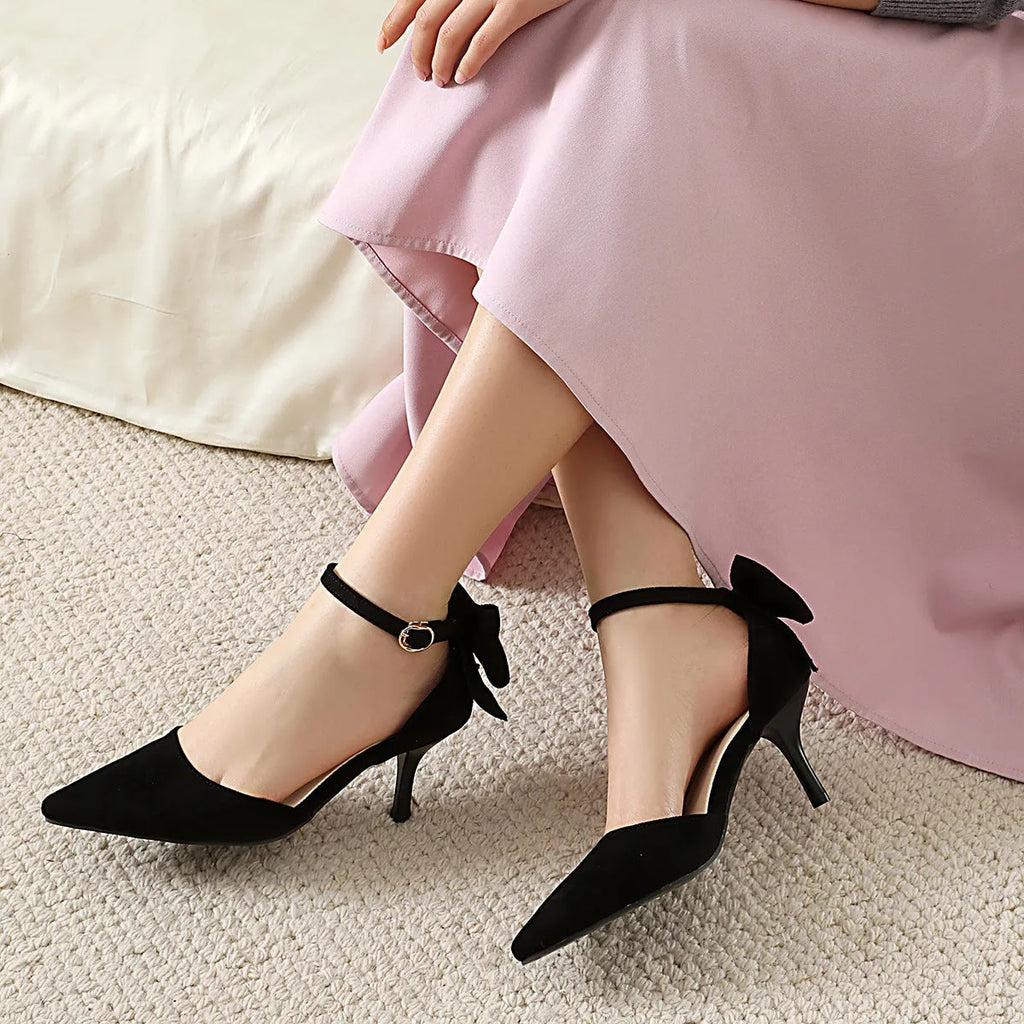 Women's black stiletto high heels with pointed toe, ankle buckle, and a bowknot detail on the suede upper.
